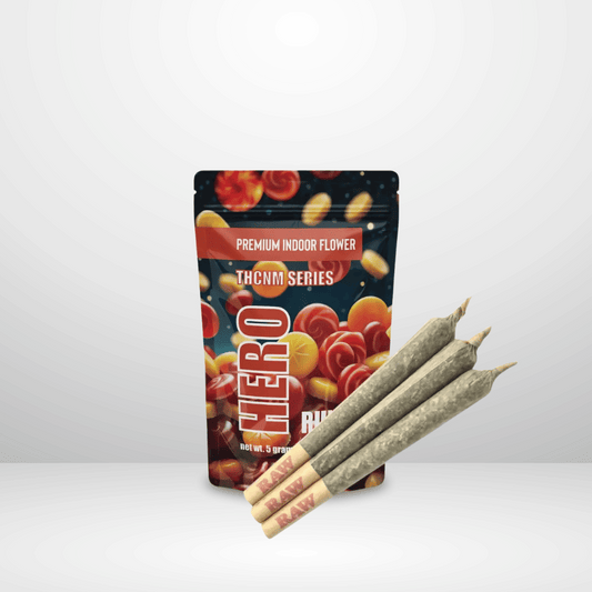En färgglad Hero-påse märkt THCNM Series Premium Indoor Flower står upprätt, med tre Hero THCNM-förrullar 50% 1st placerade framför på en vanlig, ljus bakgrund.