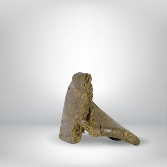 En 5 g stor bit av Partybolagets Dark Charas THCA Hash (31%), solid och brun med grova kanter, står upprätt mot en vanlig ljusgrå bakgrund.