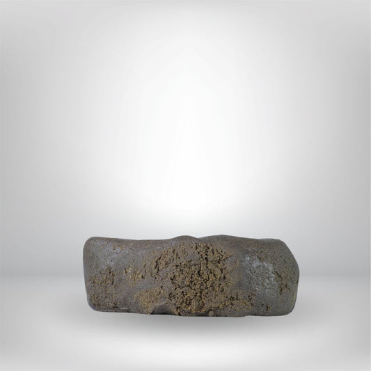 En ungefär rektangulär, mörkbrun Light Charas THCA 5g 30% - Hash från Partybolaget med en ojämn, strukturerad yta, mot en vit bakgrund med dämpad belysning. Premiumhantverk och lagligt i Sverige.